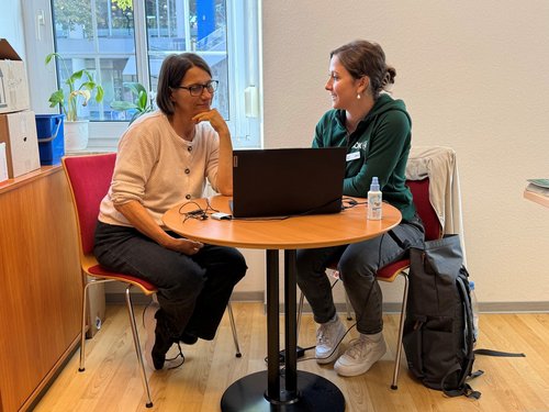 Zwei Frauen sitzen sich an einem runden Holztisch gegenüber und führen ein Gespräch. Ein geöffneter Laptop steht zwischen ihnen. Die Frau links trägt eine Brille und einen hellen Pullover, die Frau rechts einen grünen Hoodie. Sie schauen sich an.