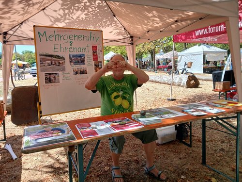 Ein Mann in einem grünen T-Shirt steht unter einem Zelt und bildet mit den Händen einen Bilderrahmen. Hinter ihm steht ein Whiteboard mit der Aufschrift „Mehrgenerativ & Ehrenamt“. Vor ihm befindet sich ein Tisch mit Informationsbroschüren.