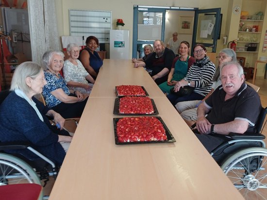 Eine Gruppe von älteren Menschen und Betreuern sitzt um einen langen Tisch in einem hellen Raum. In der Mitte stehen drei große Backbleche mit rotem Belag, vermutlich Pizzas. Die Personen lächeln und wirken gesellig; einige sitzen im Rollstuhl.