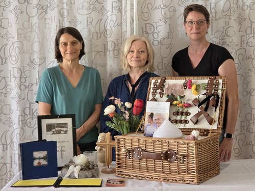Drei Frauen stehen hinter einem Tisch, auf dem ein großer geflochtener Korb mit verschiedenen kleinen Gegenständen, ein Blumenstrauß und gerahmte Bilder stehen.