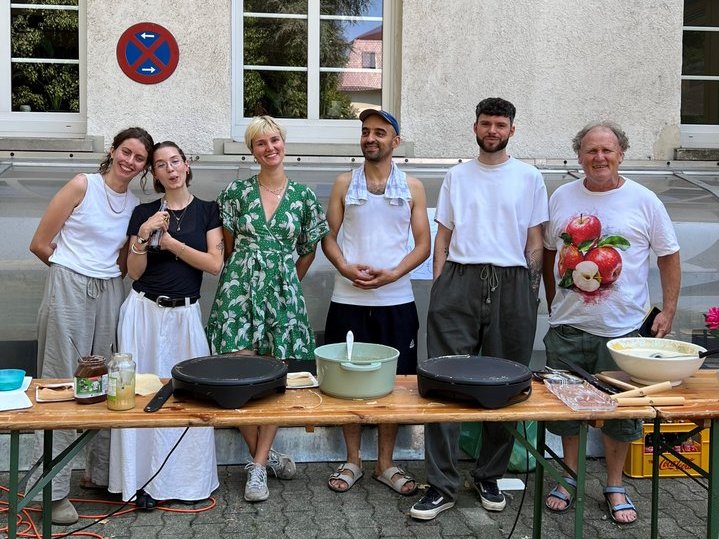 Sechs Personen stehen hinter einem langen Holztisch im Freien. Auf dem Tisch befinden sich Kochutensilien, darunter zwei große Pfannen und mehrere Schüsseln. Es wirkt wie eine Kochveranstaltung oder ein Workshop, da ein Mann zudem ein Schild mit Gemüse hält.