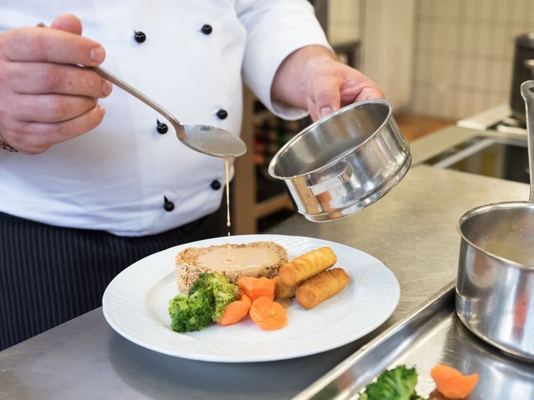 Nahaufnahme in der Küche: ein Koch gibt Soße auf einen Teller mit Fleisch und Gemüse