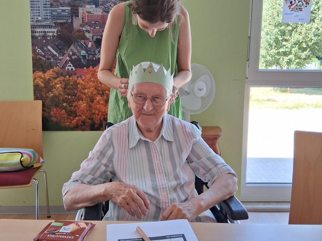 Eine ältere Dame im Rollstuhl sitzt an einem Tisch vor einem Bingo-Zettel. Eine jüngere Frau steht hinter ihr und setzt ihr einen Papierhut auf. Die ältere Dame lächelt.